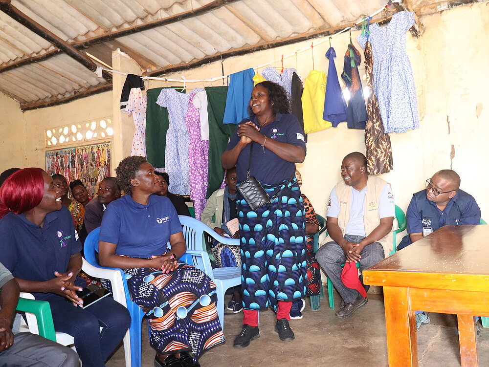 Several people are sitting on chairs in a room. A woman stands in the middle of the room and appears to be speaking. Clothes are hanging on the wall behind her.