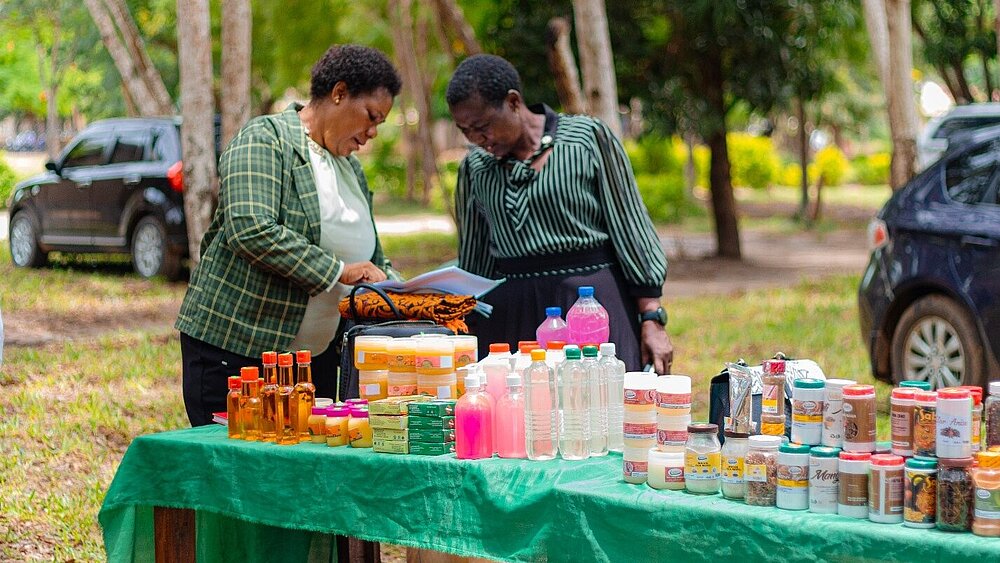 Two women stand outdoors at a table displaying various products such as bottles, jars, and packaged goods. They are looking at documents together.