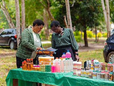 Two women stand outdoors at a table displaying various products such as bottles, jars, and packaged goods. They are looking at documents together.