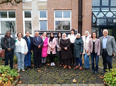 Dreizehn Männer und Frauen stehen nebeneinander in einer Reihe vor einem roten Backsteingebäude. Sie blicken lächelnd in die Kamera. 