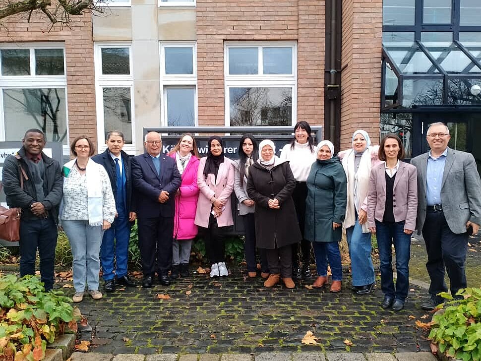 Thirteen men and women stand side by side in a row in front of a red brick building. They smile at the camera. 