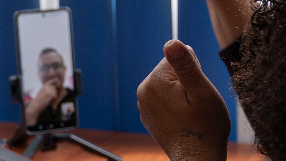 Im Vordergrund sind zwei erhoben Daumen zu sehen, die einer Person im Hintergrund gewidmet sind. Die Person ist via Videoanruf auf einem Smartphone zu sehen.