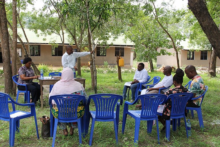 A small group of people sits outdoors on blue plastic chairs arranged in a circle. One person stands and speaks to the group