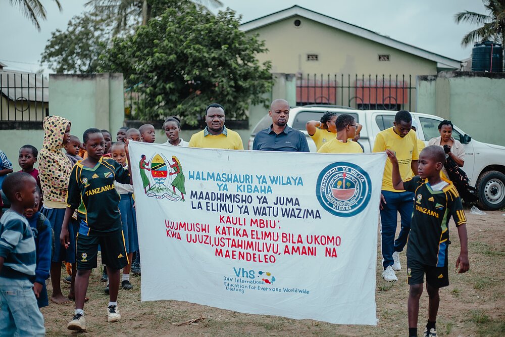 A group of children and adults hold a large banner together. On the left and right of the banner are the Tanzanian coat of arms and another logo of the Kibaha region. At the bottom is the logo of DVV International with the slogan ‘Education for Everyone. Worldwide. Lifelong.’ In the background are trees, buildings and a few other people.