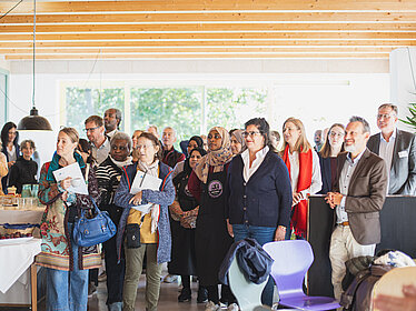 Eine Gruppe von Menschen steht in einem hellen Raum. Sie blicken interessiert nach links, auf eine Person außerhalb des Bildrandes. Im Vordergrund sind Stühle und ein Tisch mit Obst und Gemüse.
