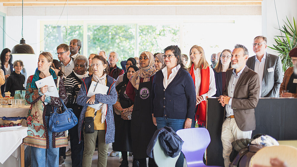Eine Gruppe von Menschen steht in einem hellen Raum. Sie blicken interessiert nach links, auf eine Person außerhalb des Bildrandes. Im Vordergrund sind Stühle und ein Tisch mit Obst und Gemüse.