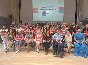Several people are in an auditorium in front of a projected image titled "Summer School, Empower Democracy." The people in the front row are seated on chairs. Everyone is holding a red envelope.