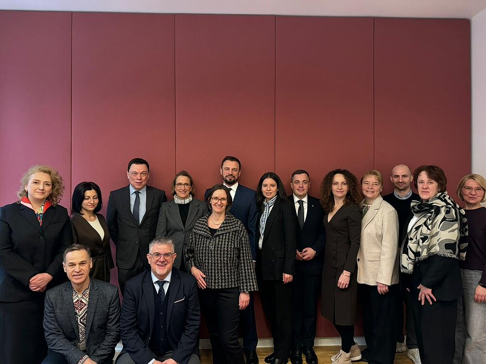Fifteen women and men stand closely together in front of a red wall. They are dressed in formal attire and are looking at the camera.