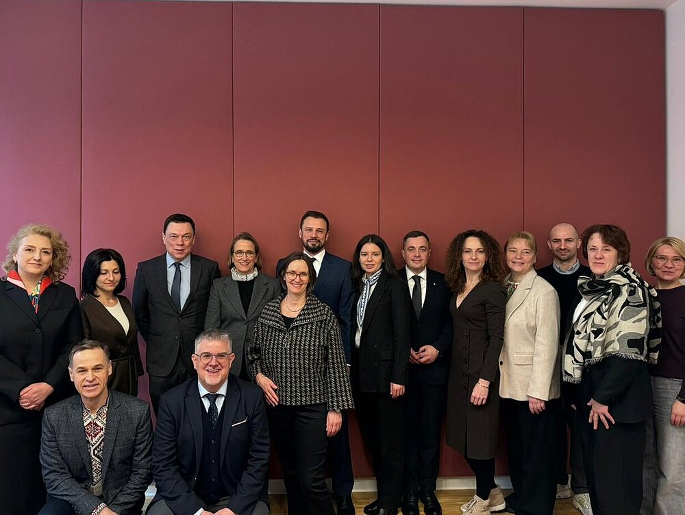Fifteen women and men stand closely together in front of a red wall. They are dressed in formal attire and are looking at the camera.