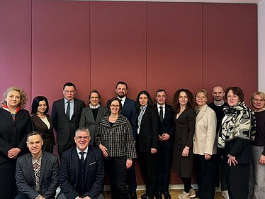 Fifteen women and men stand closely together in front of a red wall. They are dressed in formal attire and are looking at the camera.