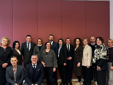 Fünfzehn Frauen und Männer stehen nebeneinander vor einer rötlichen Wand. Sie sind formell gekleidet und blicken in die Kamera.
