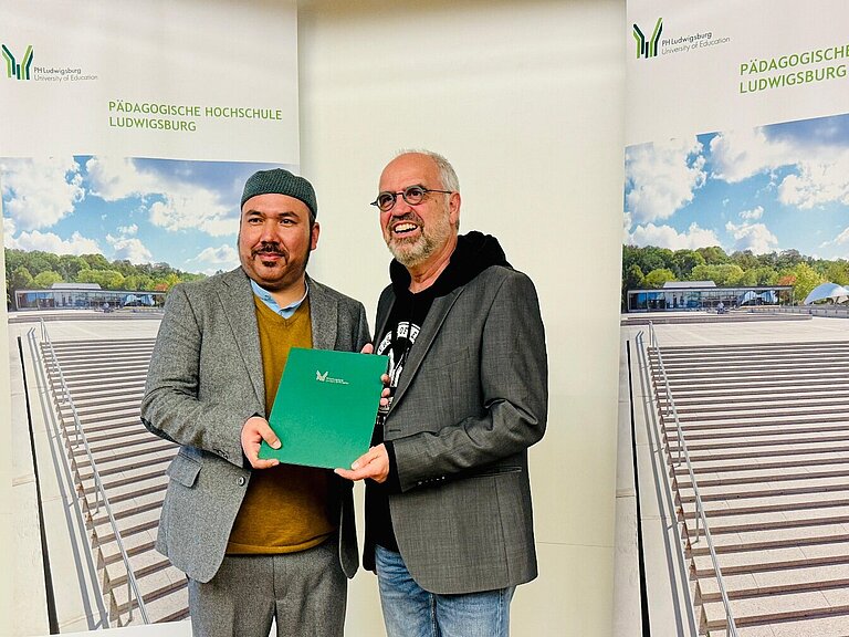 Zwei Männer stehen vor einem Banner der Pädagogischen Hochschule Ludwigsburg und halten gemeinsam eine grüne Mappe in den Händen.