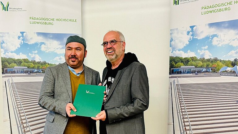 Two men stand in front of roll-up banners of the Ludwigsburg University of Education, holding a green certificate together.