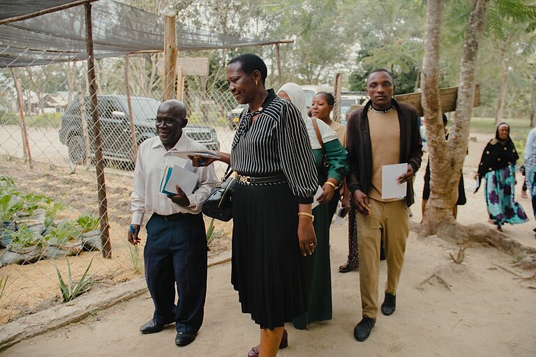 Several people walk together along a path. They are in a rural setting with trees, a fence, and a vegetable garden visible in the background.