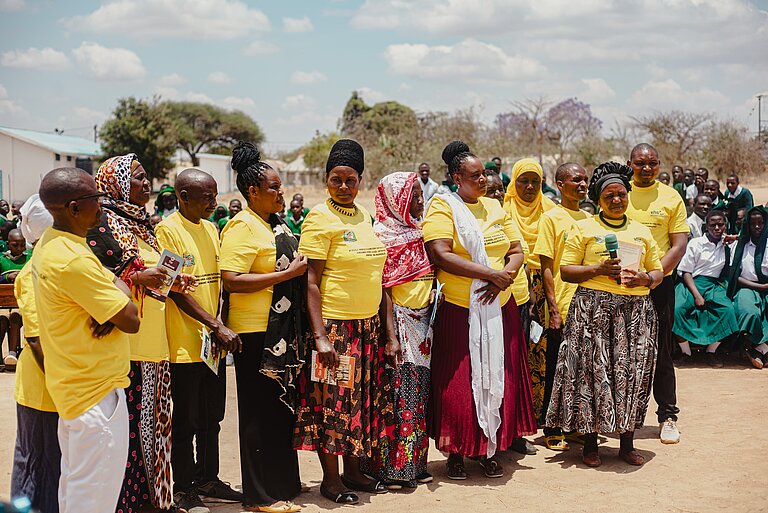 A group of people, mostly women, is standing together outdoors, wearing bright yellow T-shirts with writing on them, possibly related to an event or awareness campaign. Some of the women are wearing traditional African clothing and colorful headscarves, while one person holds a microphone, appearing to address the group or audience. Behind them, people, including children in school uniforms, are gathered, watching the scene. 