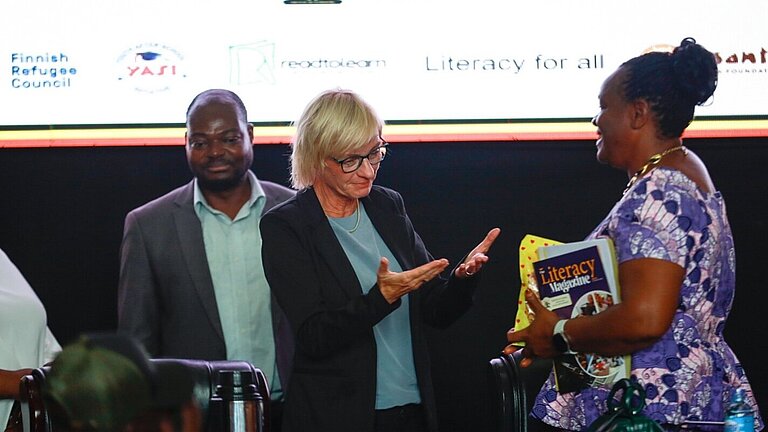 A woman opens her hands toward another lady who is holding books in her arms. A man is standing in the background.
