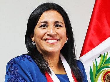 A woman stands against a light background, smiling at the camera. She is wearing a blue dress and a red-and-white sash with a medal.