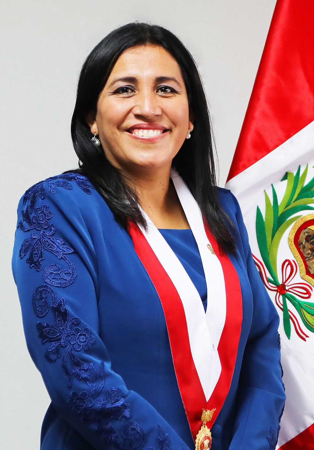 A woman stands against a light background, smiling at the camera. She is wearing a blue dress and a red-and-white sash with a medal.