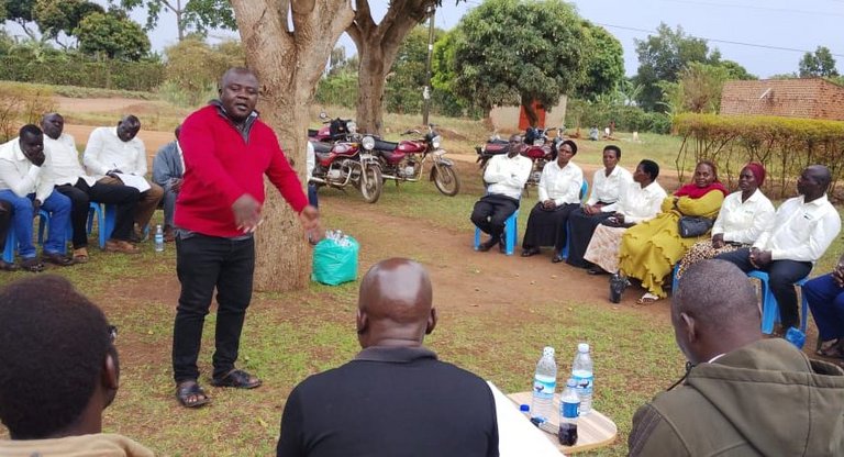 A group of people is sitting in a circle outside. A man in the middle appears to be talking and gesturing.
