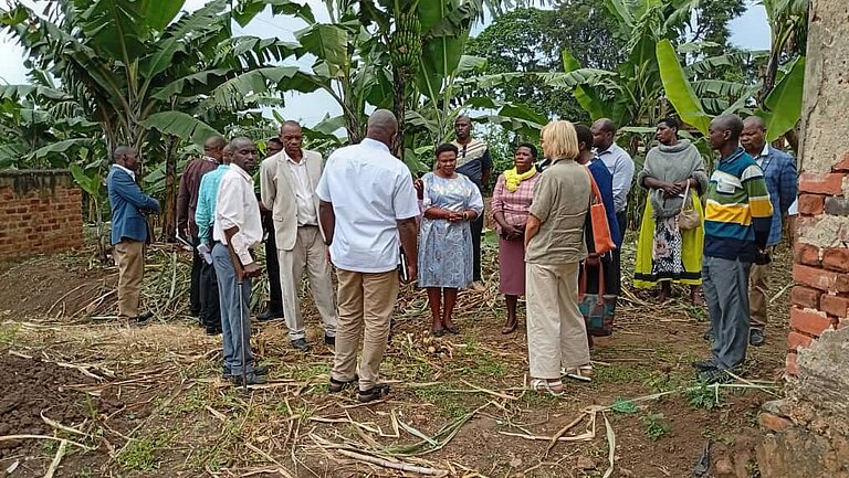 A group of people is standing outside. Most people are facing the camera, one man and one woman are turned towards the group. In the background are banana trees.