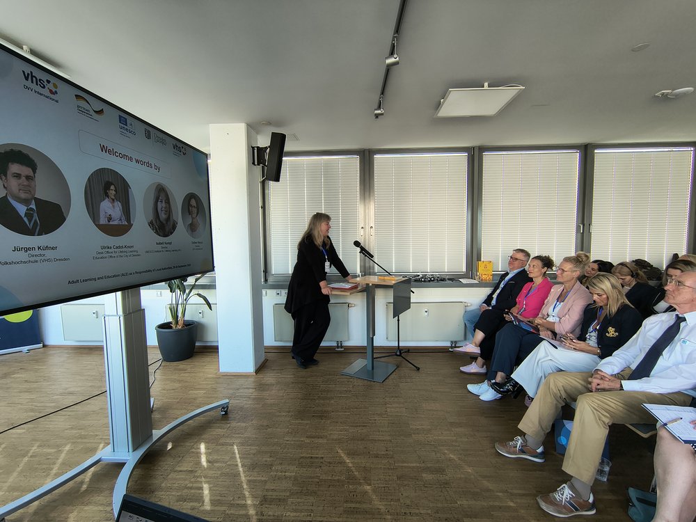 Several people are sitting on chairs in front of a smartboard. Next to the smartboard, a woman is standing at a lectern and speaking.
