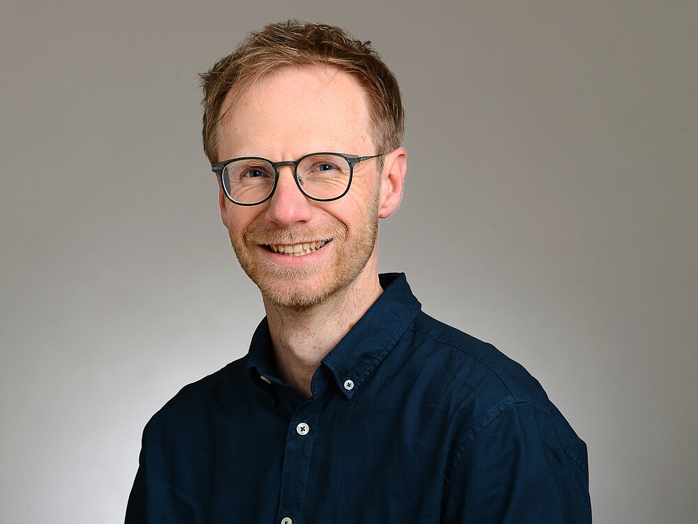 Porträtfoto von Stefan Markov, er lächelt in die Kamera.. Er trägt kurzes Haar und ein dunkelblaues Hemd mit Knöpfen vor neutralem Hintergrund.