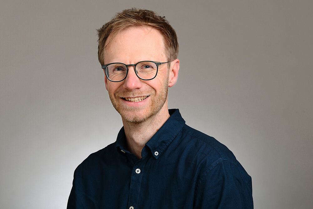 Porträtfoto von Stefan Markov, er lächelt in die Kamera.. Er trägt kurzes Haar und ein dunkelblaues Hemd mit Knöpfen vor neutralem Hintergrund.