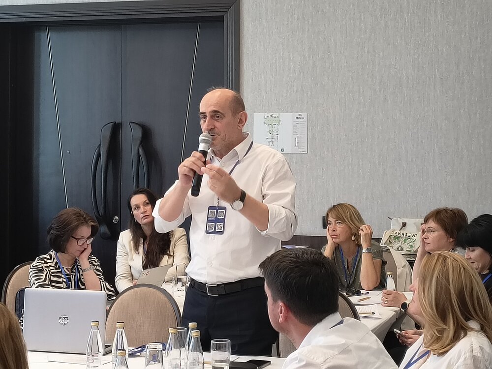 A man is standing in a conference room, speaking into a microphone. Other participants are seated around him at conference tables, listening to his presentation.