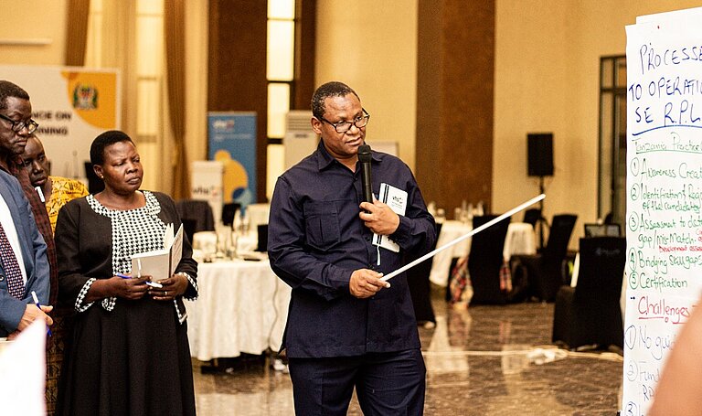 A man is standing in front of a poster, speaking into a microphone and holding a pointer in his other hand. A few people stand behind him and follow his presentation. The people find themselves in a formal function room. 