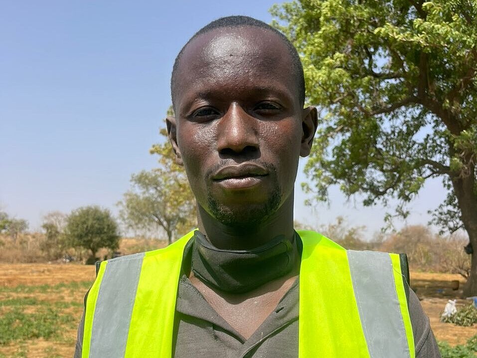 A young man stands outdoors in a field, looking directly at the camera. He is wearing a bright yellow safety vest.