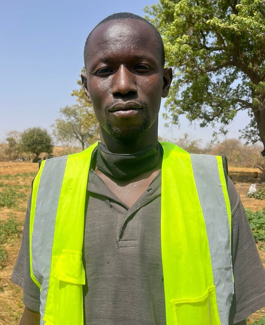 A young man stands outdoors in a field, looking directly at the camera. He is wearing a bright yellow safety vest.