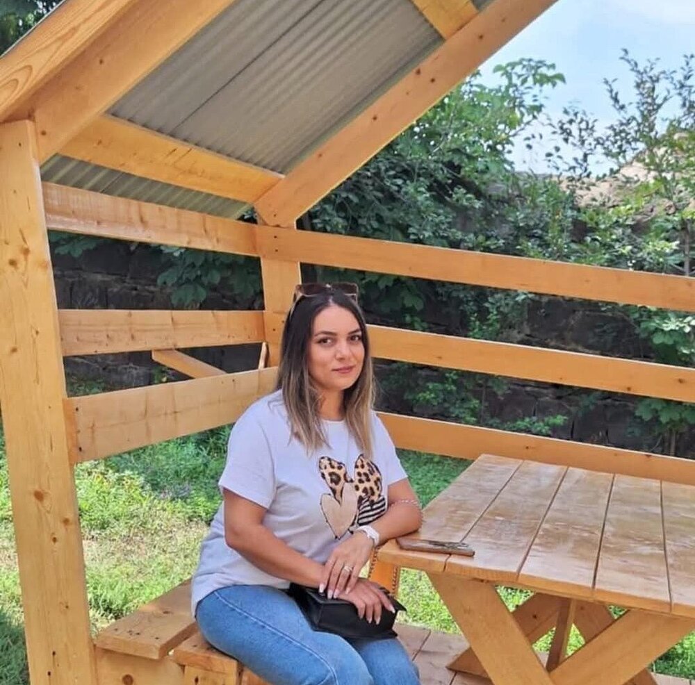 A woman is sitting outside on a bench at a table, holding her handbag on her lap.