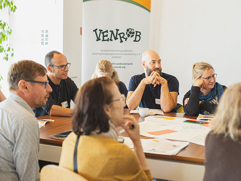 Personen sitzen an einem Tisch und blicken nach rechts. Im Hintergrund ein VENROB Banner.