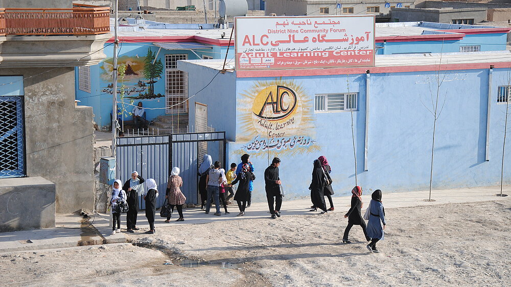 Several women wearing headscarves and two boys are standing in front of a light blue building. On the roof of the building is a sign that reads “Adult Learning Center”