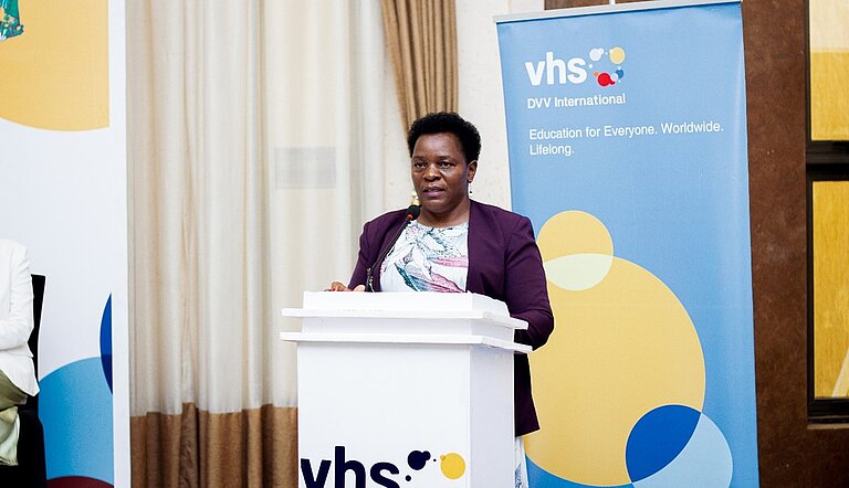 In a formal event room, a woman stands at a lectern, addressing the audience. Behind her, a banner bearing the DVV International logo.
