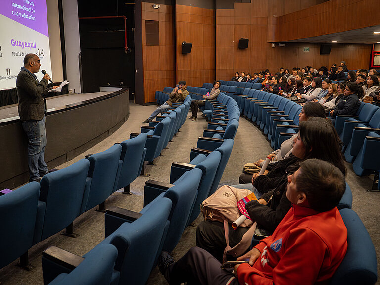 Several people are sitting in a cinema auditorium. A man is standing at the front of the stage speaking into a microphone.