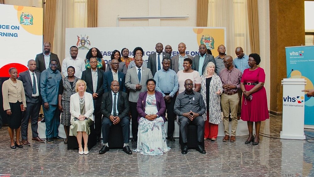 Group photo of participants at a conference. The people are standing and sitting in several rows in a formal event room. Banners with the logos of vhs DVV International and a government agency can be seen in the background. A lectern with vhs branding is on the right of the picture. The participants are wearing formal clothes, some are smiling while others are looking neutrally into the camera.