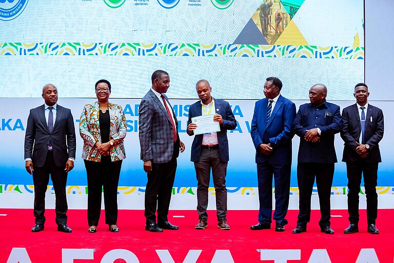 Seven people in suits are standing next to each other, the one in the middle holding a certificate.